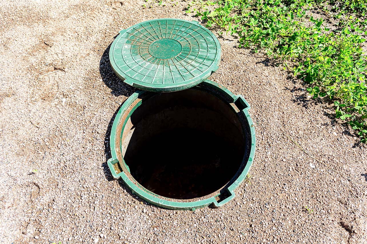 The opening of a green underground septic tank with the green lid off and laid to the side of the opened tank.