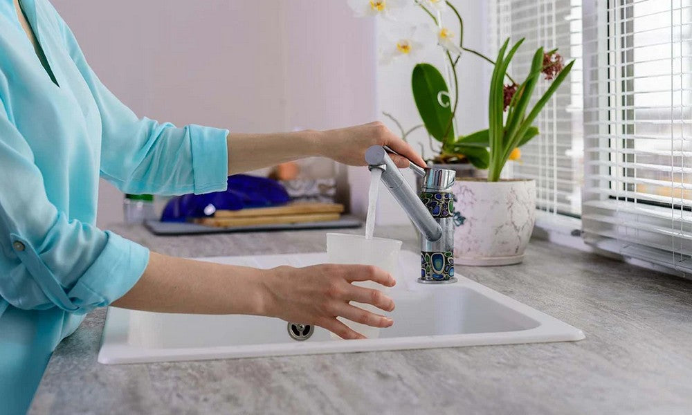 A woman in a blue shirt at a kitchen sink turning on the faucet to fill a glass with water.