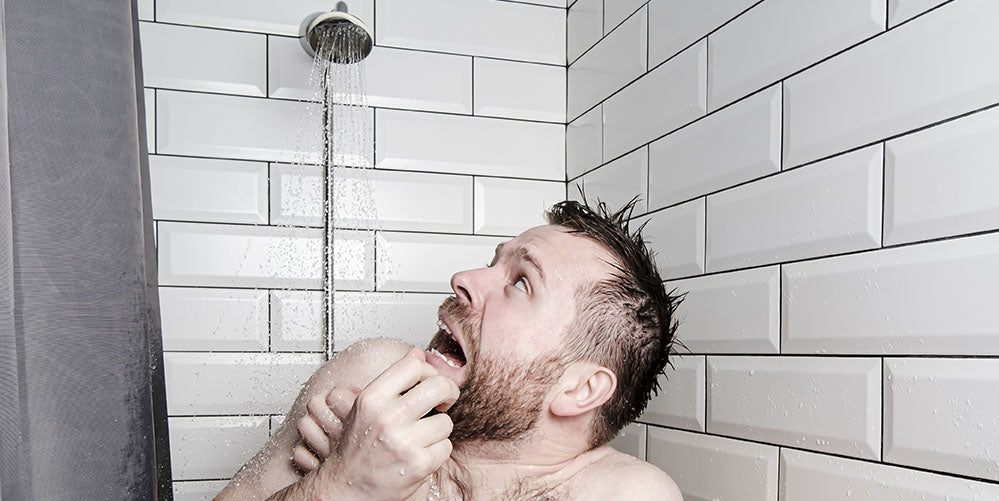 A man in a tile shower looking up at the cold water coming from the shower head.