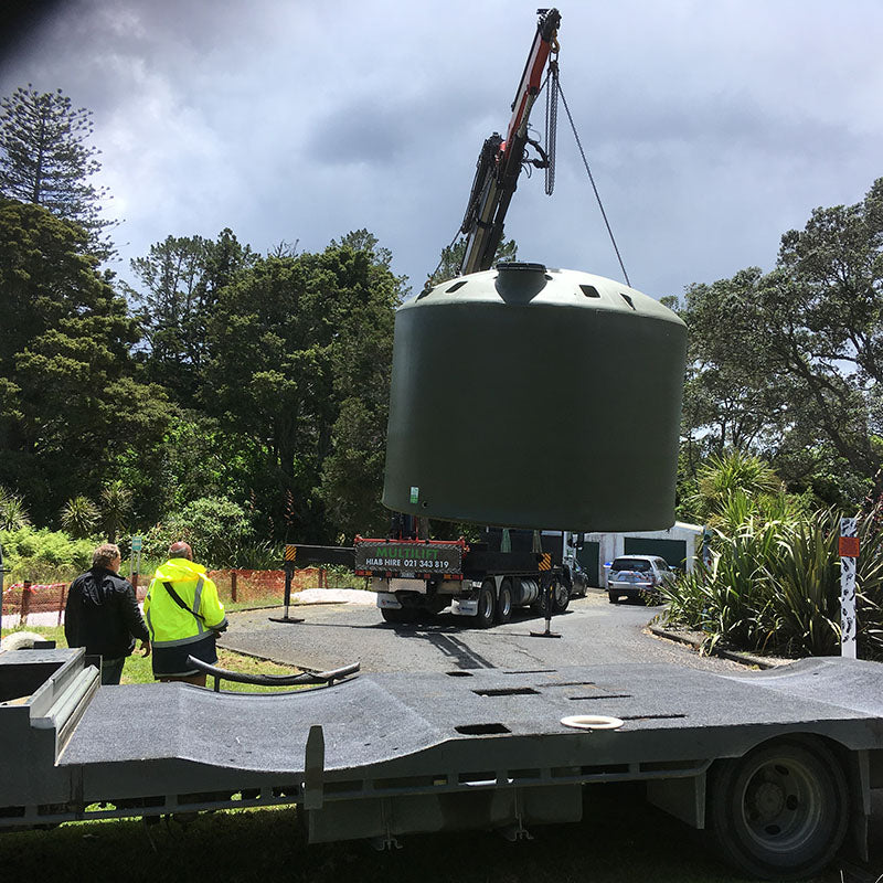 A crane lifting a large green water tank from a truck bed while two high visability vested people look on.